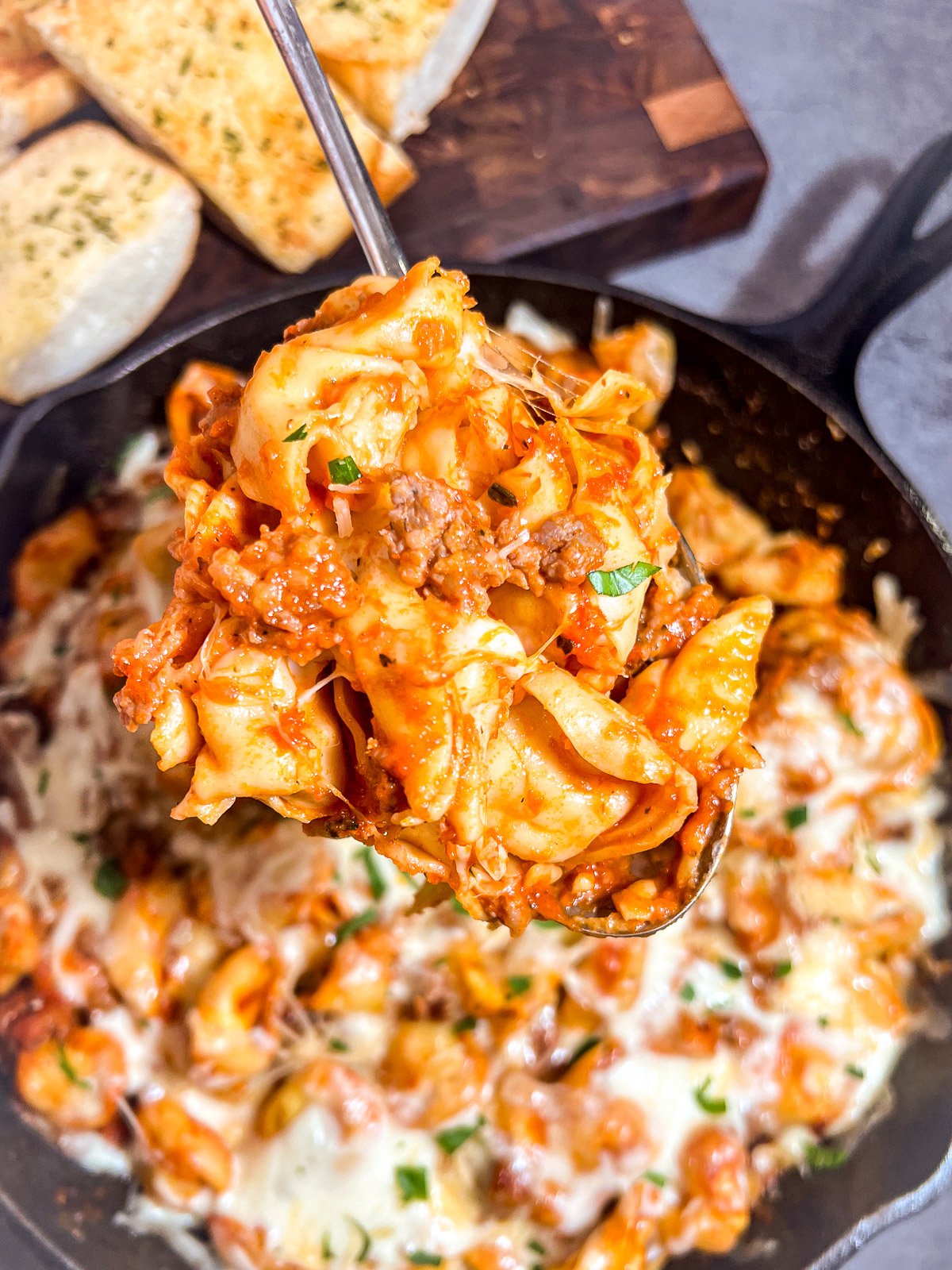 Spoon scooping baked tortellini with sausage and melted cheese from cast iron skillet