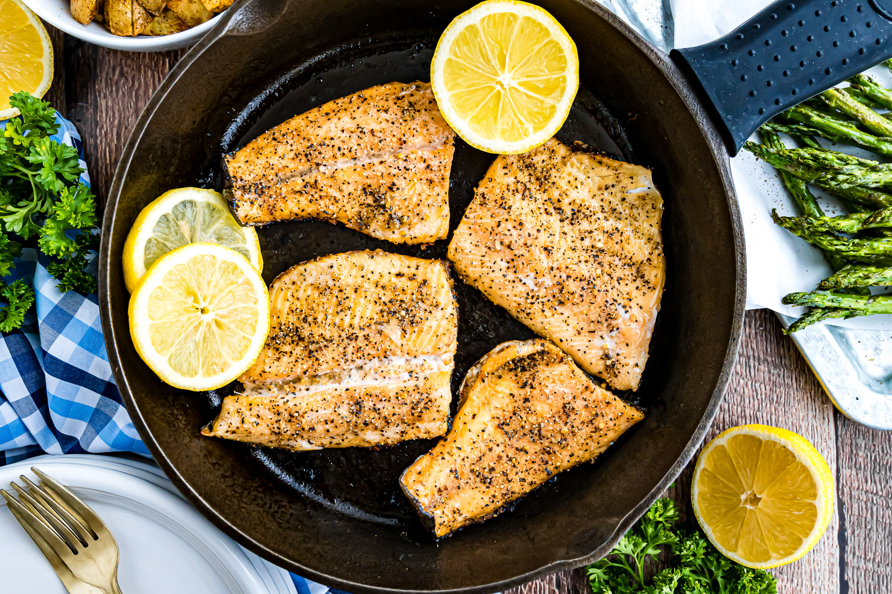 Overhead view of grilled rainbow trout fillets in a cast iron skillet with lemon slices and asparagus