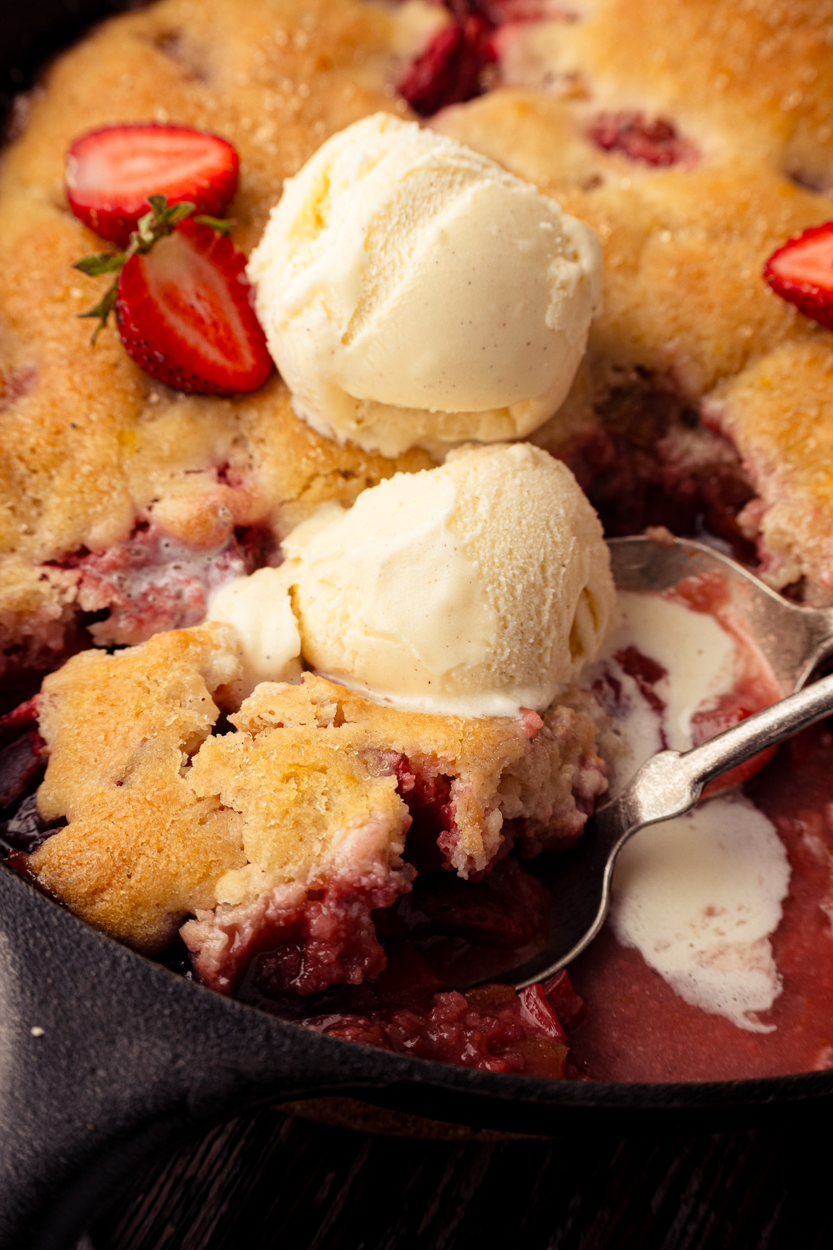 Skillet strawberry rhubarb cobbler with vanilla ice cream scoops and fresh strawberry slices in a cast iron skillet