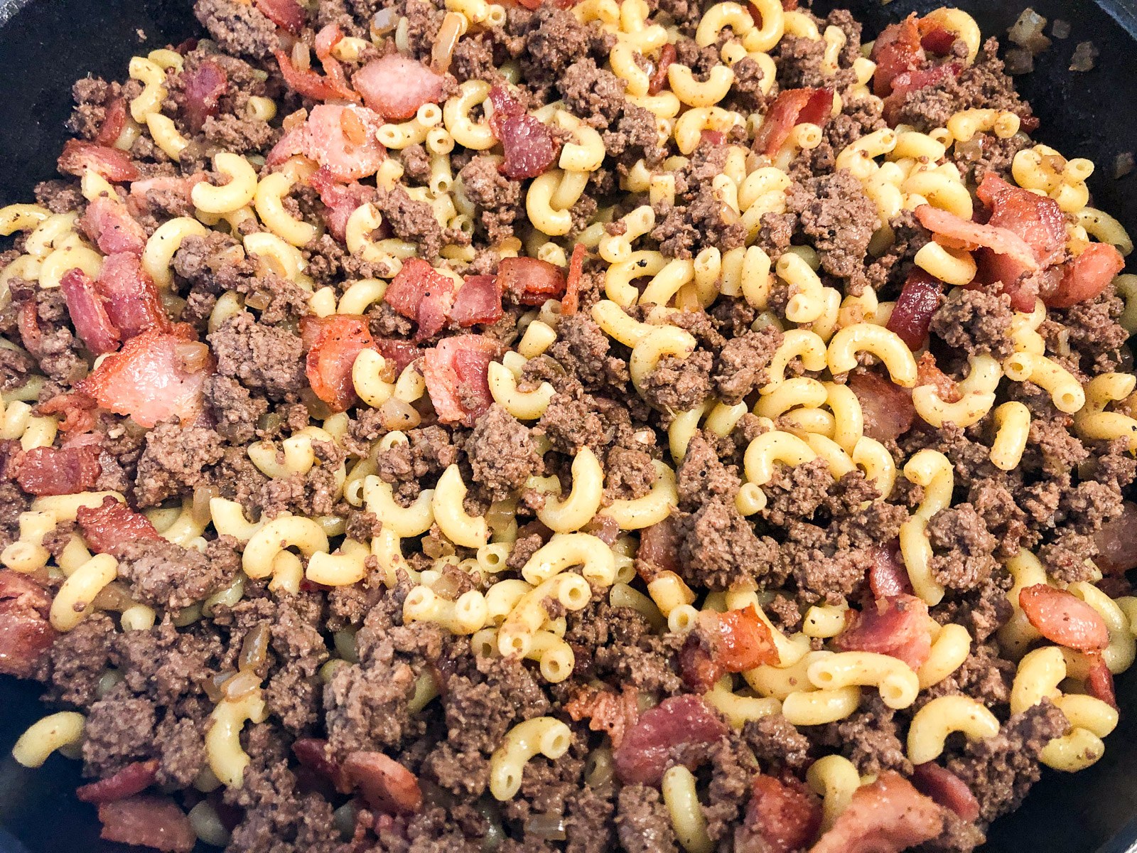 Browned ground beef with bacon and seasonings in a skillet.