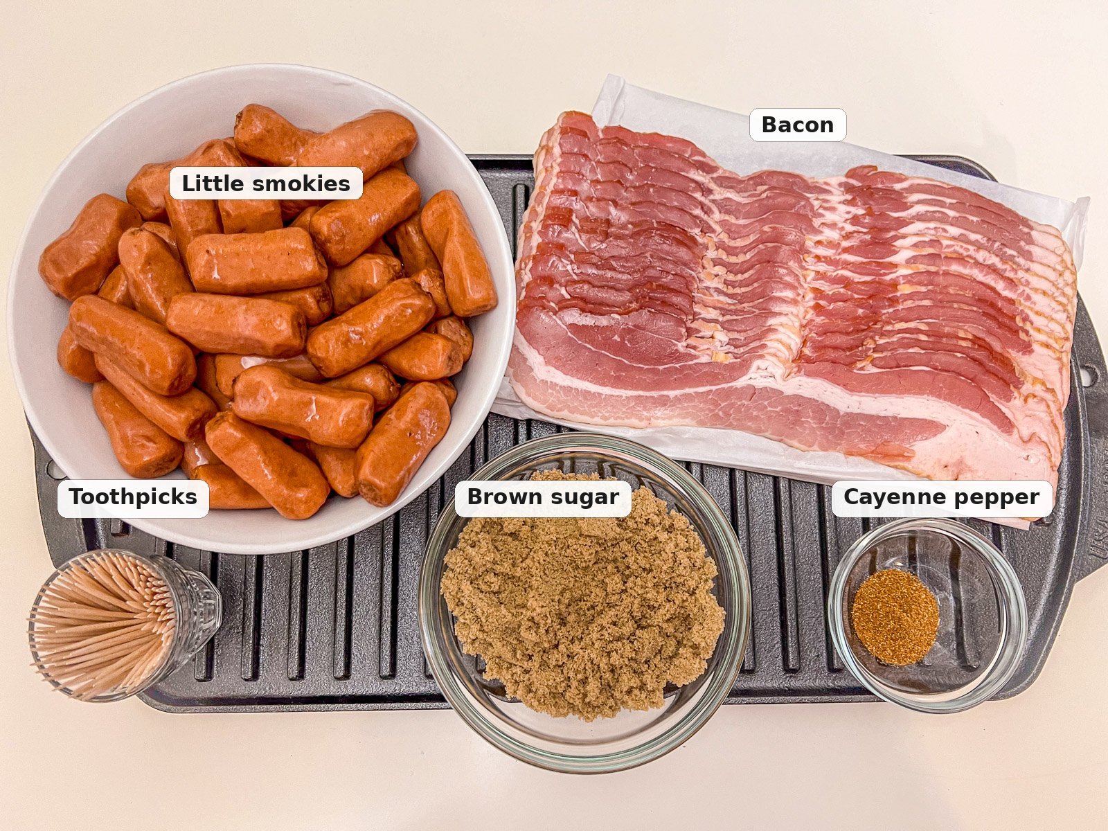 Ingredients for bacon wrapped smokies: little smokies, bacon, brown sugar, cayenne pepper, and toothpicks on a cast iron pan.
