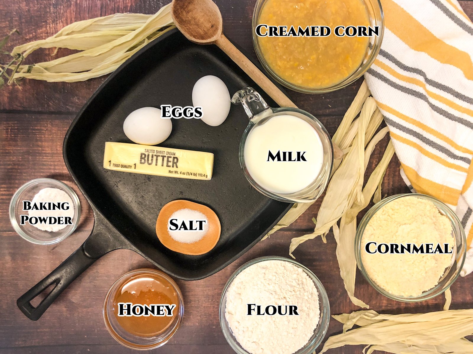Labeled ingredients for cornbread with creamed corn including cornmeal, flour, creamed corn, milk, eggs, butter, honey, baking powder, and salt.