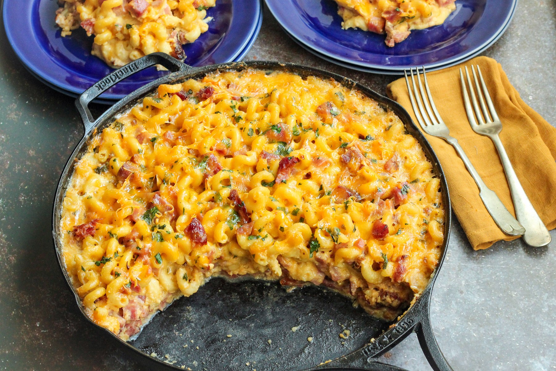 Baked mac and cheese with ham in a cast iron skillet with a serving on a blue plate