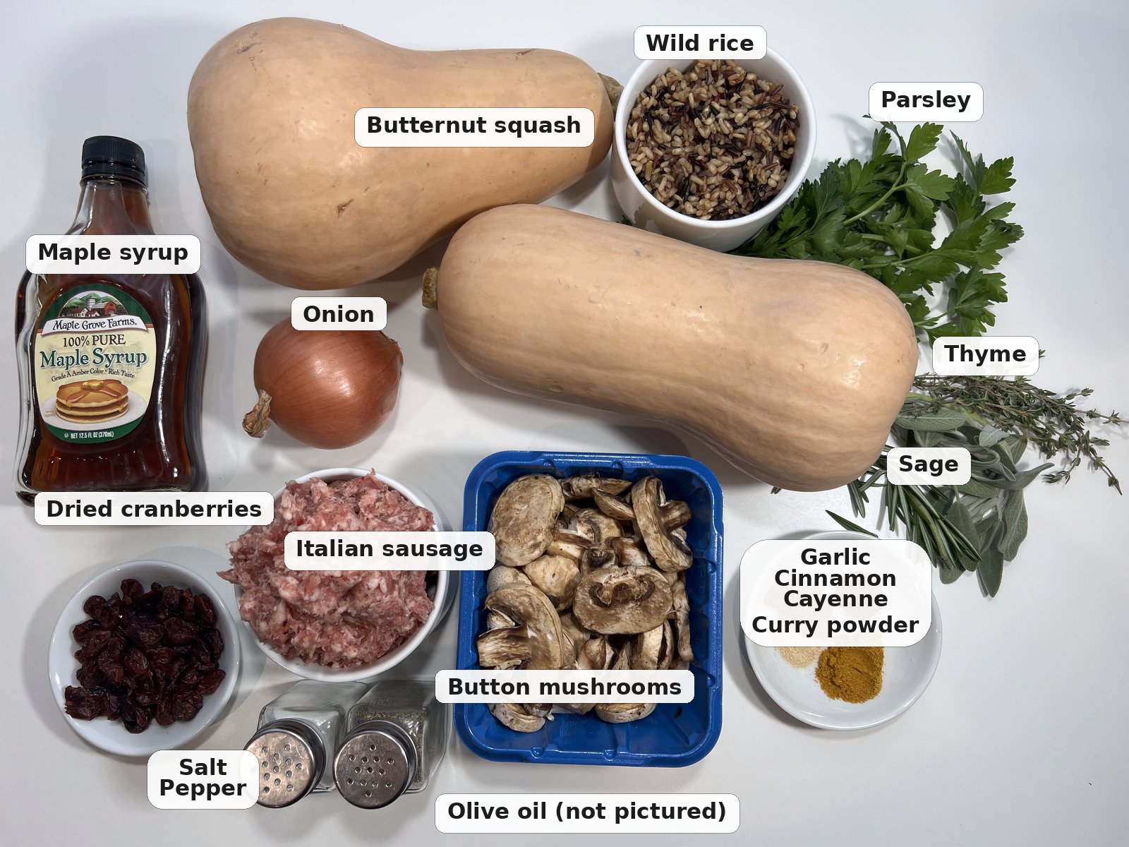 Labeled ingredients for sausage stuffed butternut squash: butternut squash, wild rice, Italian sausage, button mushrooms, dried cranberries, onion, maple syrup, parsley, thyme, sage, and spices.