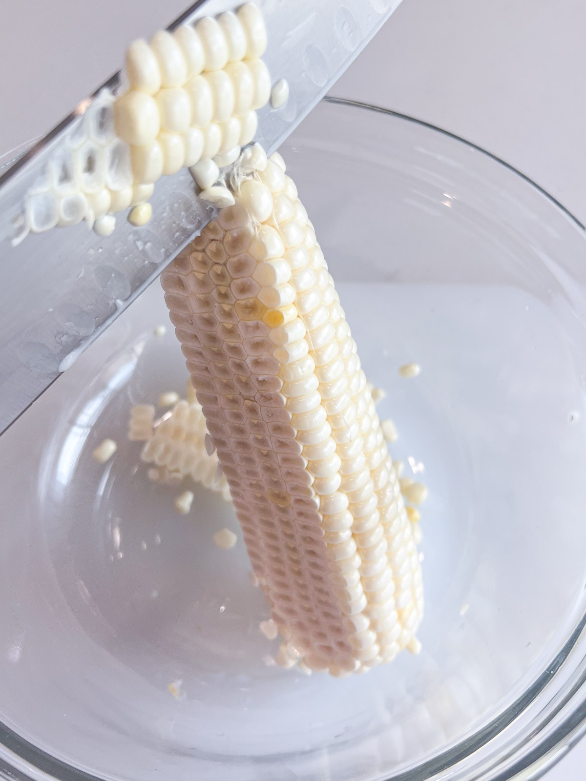 Slicing fresh sweet corn kernels off the cob into a glass bowl.