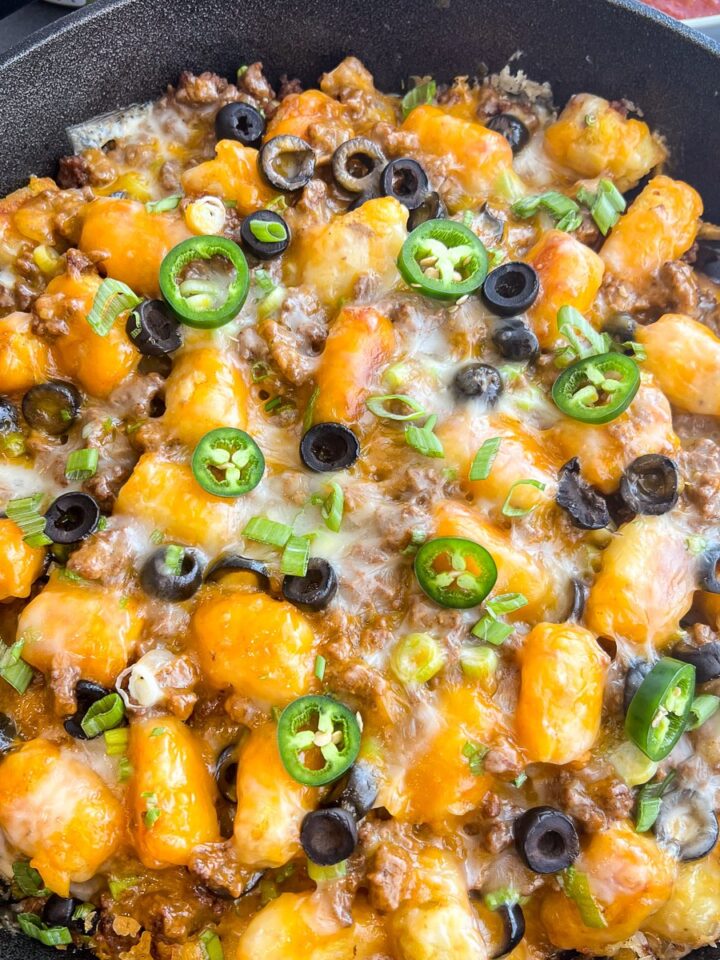 Tater tot nachos in a cast iron skillet topped with cheese, jalapenos, and black olives.