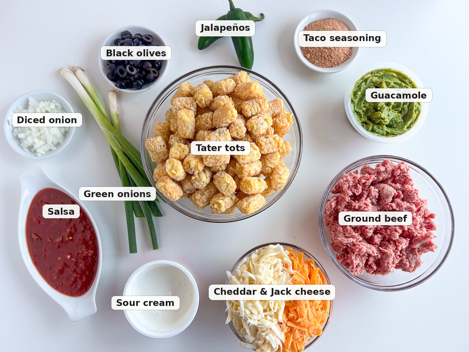 Labeled ingredients for tater tot nachos: tater tots, ground beef, taco seasoning, cheddar and Jack cheese, guacamole, salsa, sour cream, black olives, jalapenos, onion, and green onions.