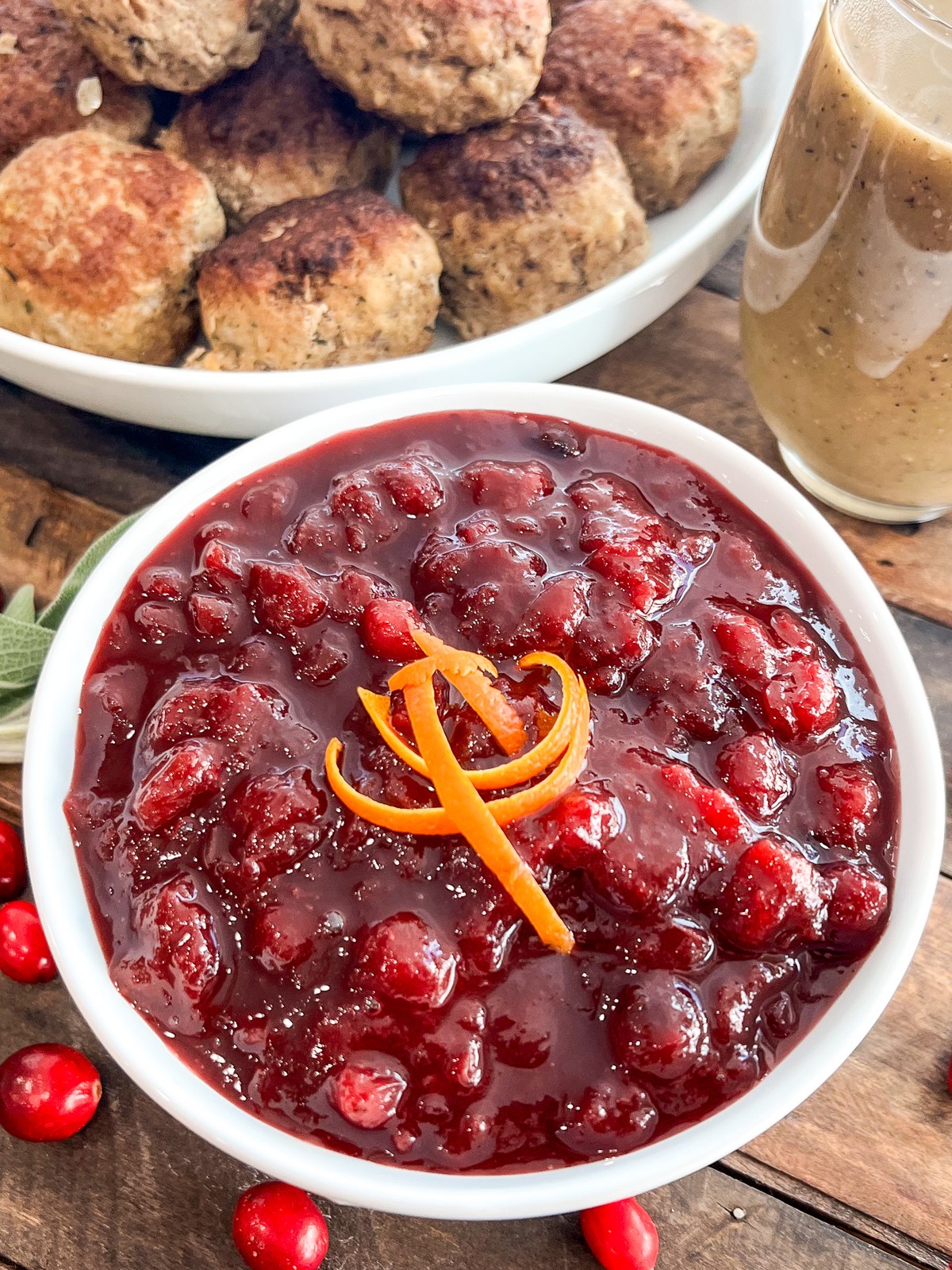 Homemade cranberry sauce garnished with orange zest, served with turkey meatballs and gravy in the background.