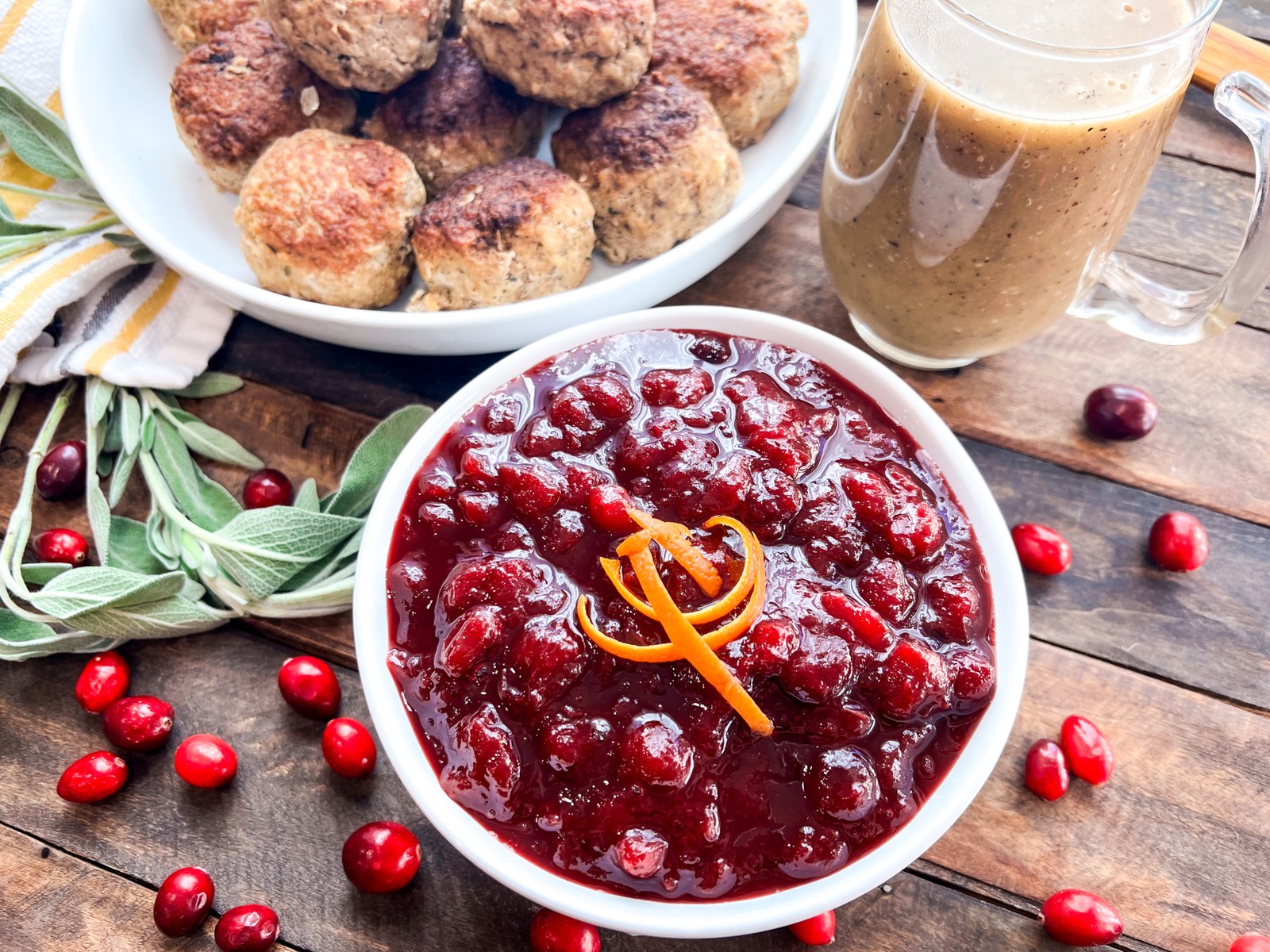 Bowl of homemade cranberry sauce with orange next to turkey meatballs, gravy, sage, and fresh cranberries.