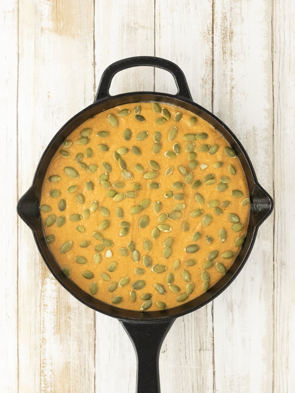 Pumpkin cornbread batter in a cast iron skillet topped with raw pumpkin seeds