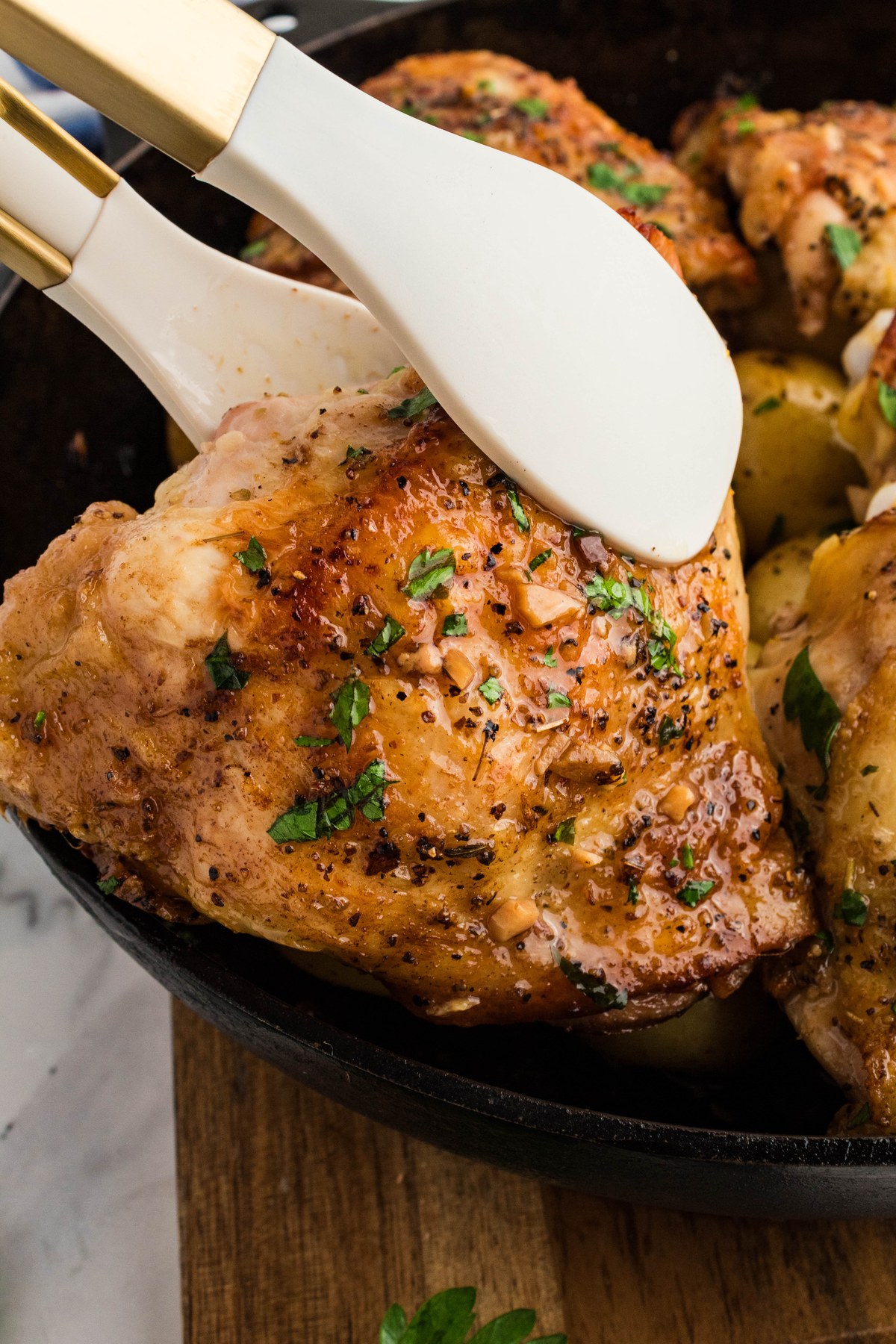 Tongs lifting a golden roasted Greek lemon chicken thigh from the skillet