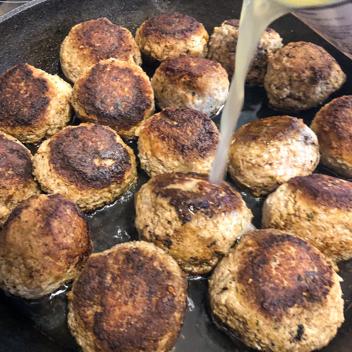 Chicken broth being poured over browned turkey and stuffing meatballs in a cast iron skillet.