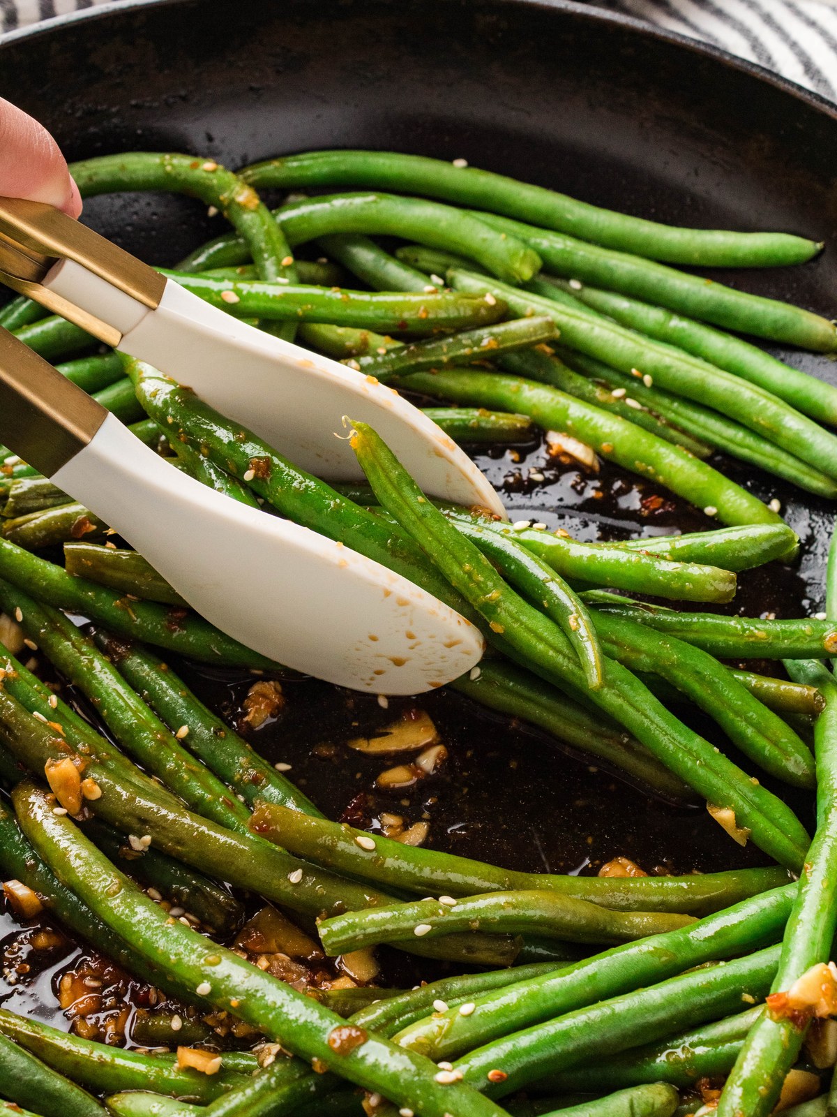 Tongs tossing Asian green beans in soy garlic sauce in a cast iron skillet
