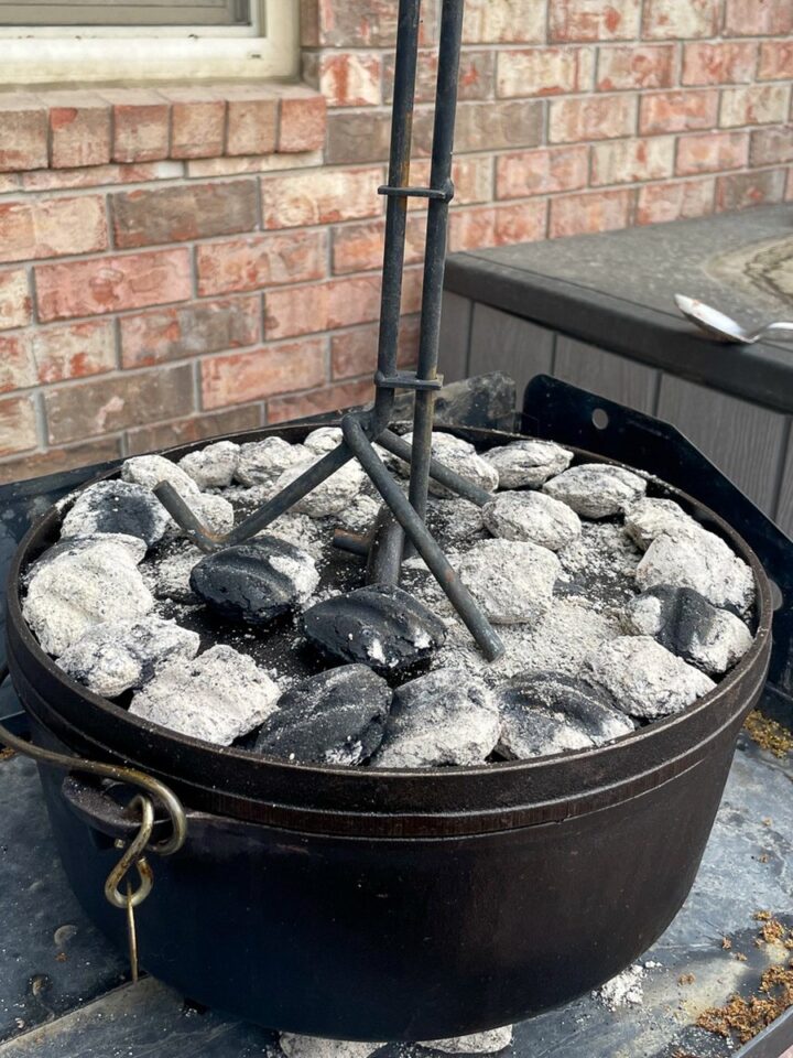A 12 inch cast iron camp oven with white ash coals arranged on the lid, ready for dutch oven camping recipes.