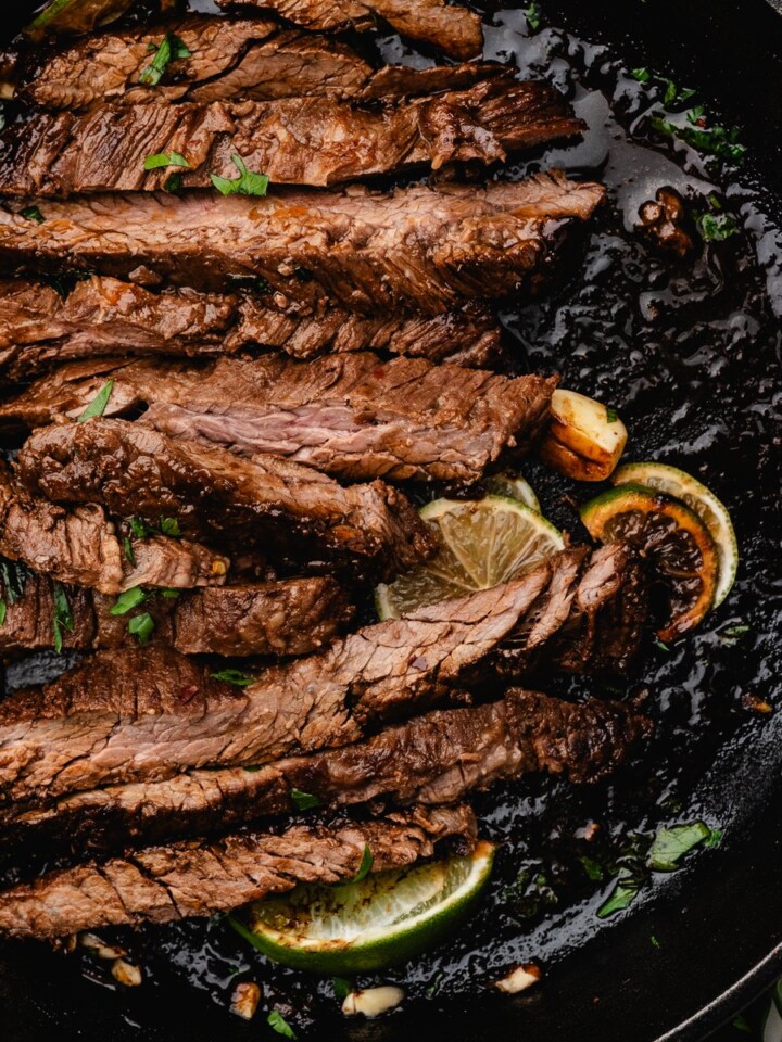 Sliced marinated steak in a cast iron skillet with lime wedges and parsley