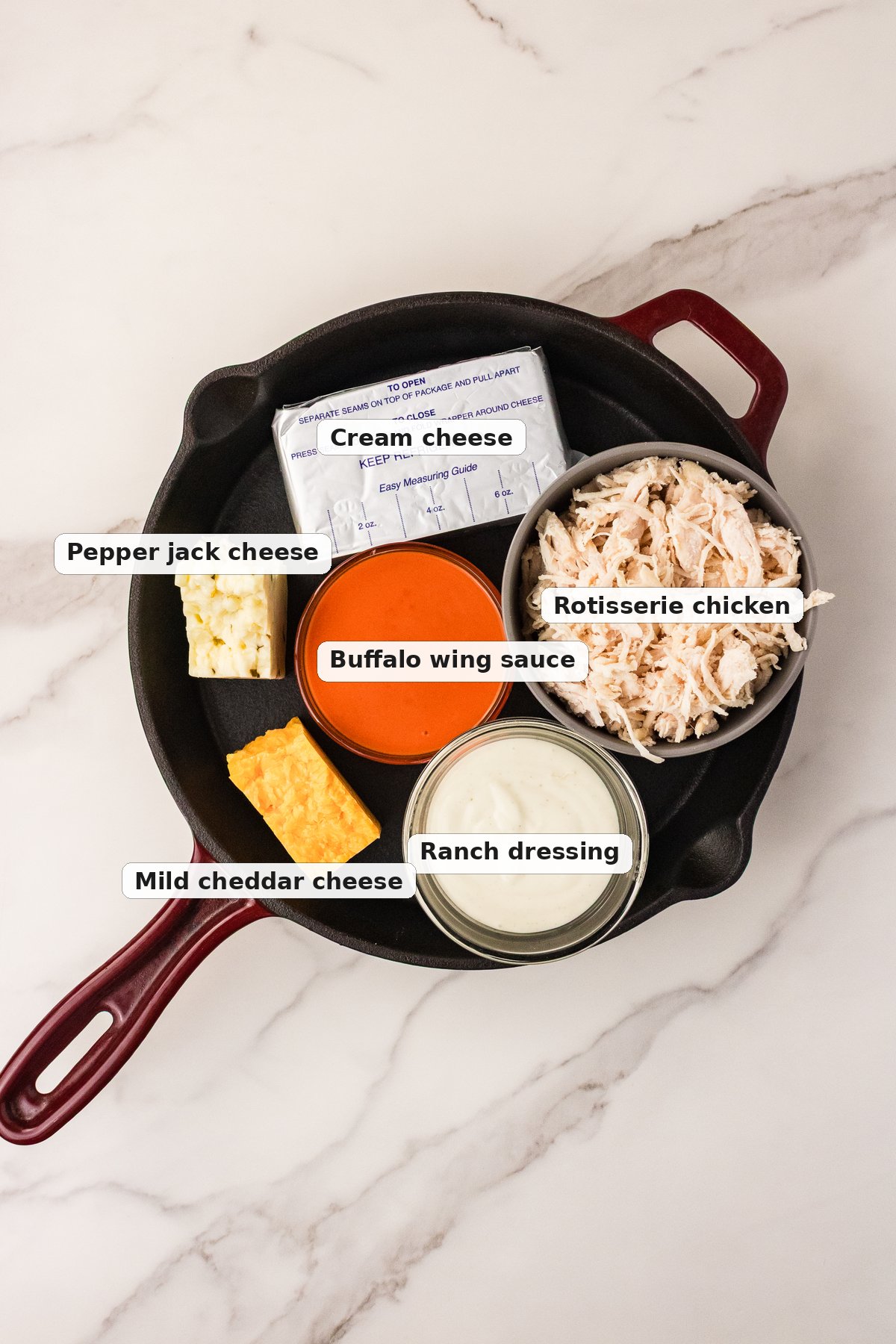 Labeled ingredients for buffalo chicken dip with rotisserie chicken in a cast iron skillet