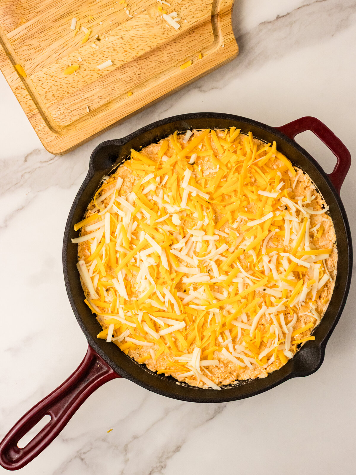 Shredded cheddar and pepper jack sprinkled over buffalo chicken dip in a skillet