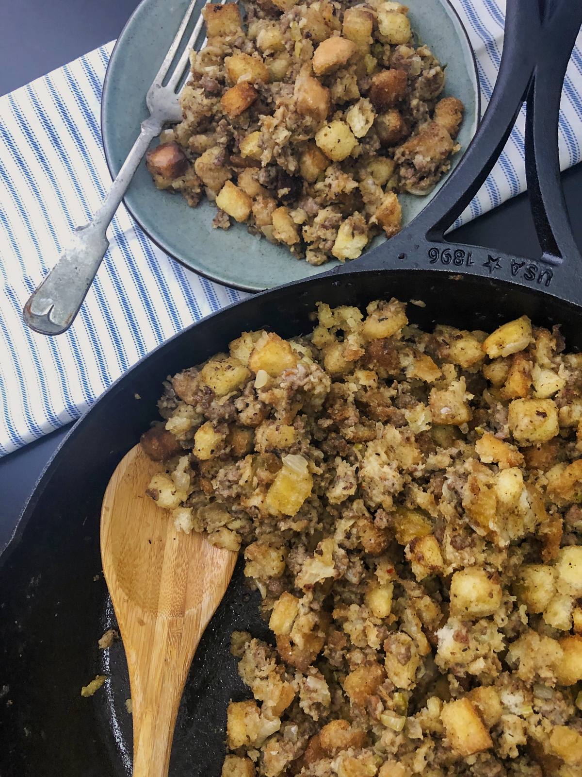 Skillet sausage stuffing in a cast iron skillet with a wooden spoon, a plated portion on a blue plate beside it.
