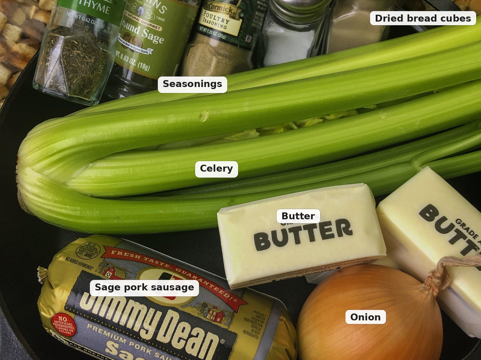 Ingredients for skillet sausage stuffing in a cast iron skillet: sage pork sausage, butter, onion, celery, dried bread cubes, thyme, rubbed sage, poultry seasoning, salt, and pepper.