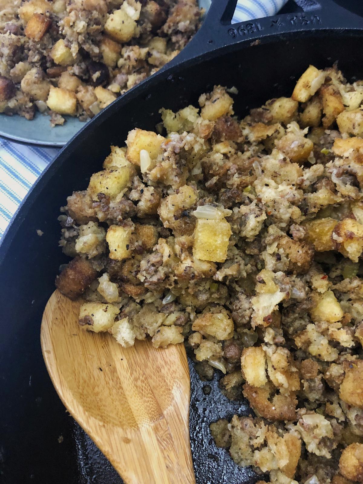 Overhead view of skillet sausage stuffing with a wooden spoon and a plated serving on a striped towel.