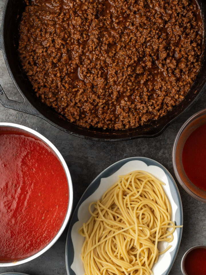 Cast iron skillet of taco meat surrounded by tomato sauce, spaghetti and shredded cheddar for taco spaghetti