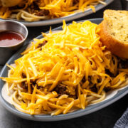 Taco spaghetti on an oval diner plate piled with shredded cheddar, garlic bread and a cup of hot sauce