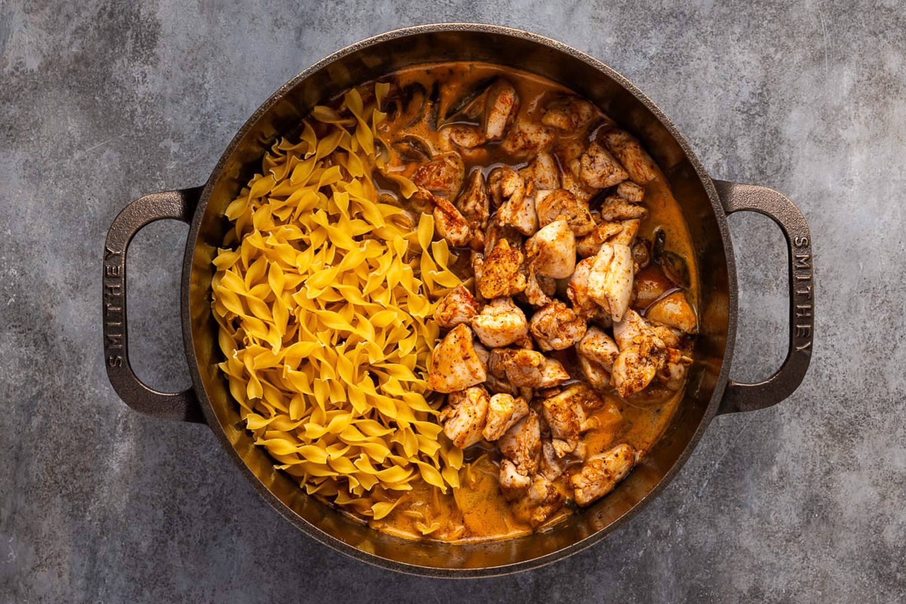 Egg noodles and seared chicken added to the simmering sauce in a Dutch oven