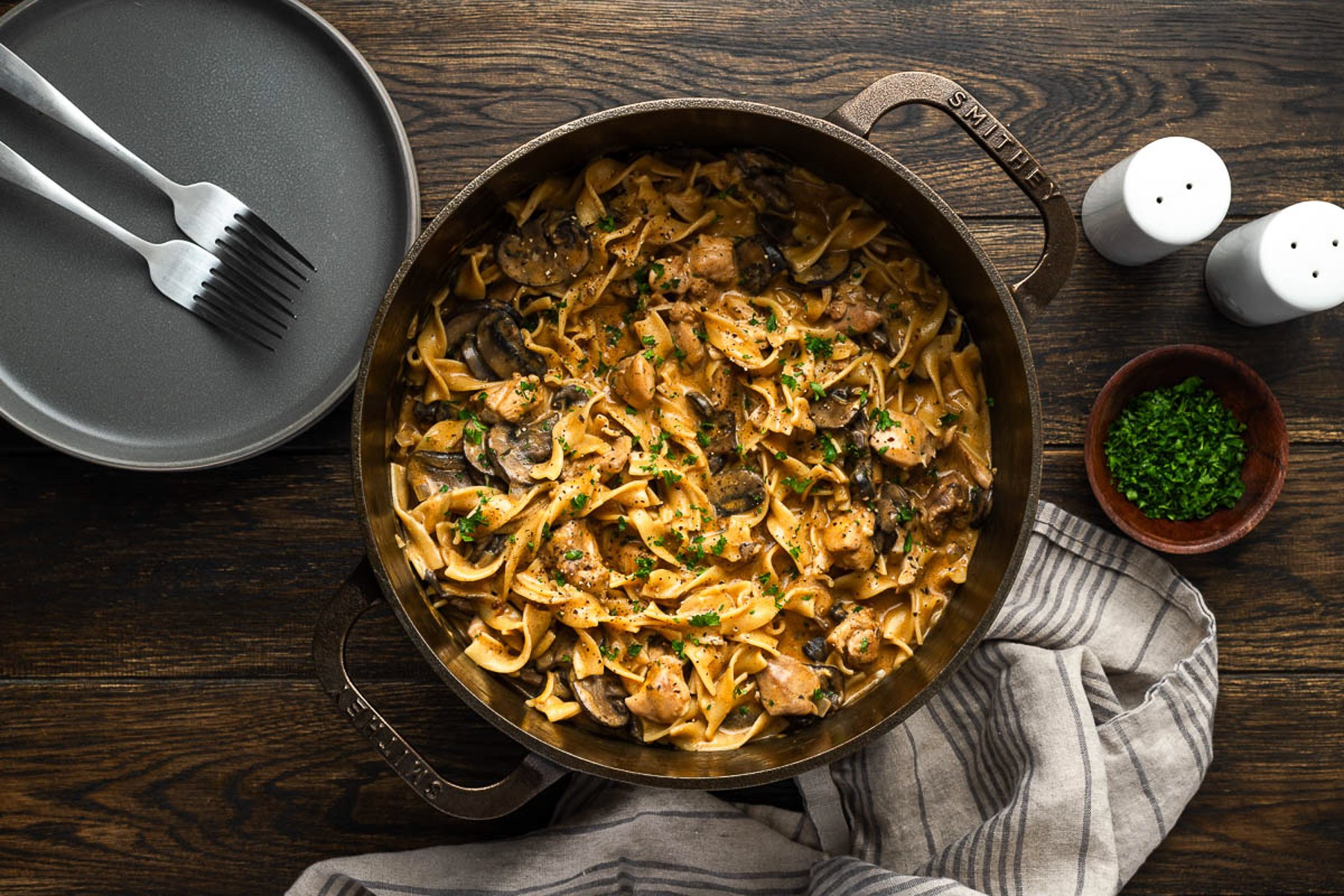 Finished chicken stroganoff in a cast iron Dutch oven with parsley, plates and forks beside it