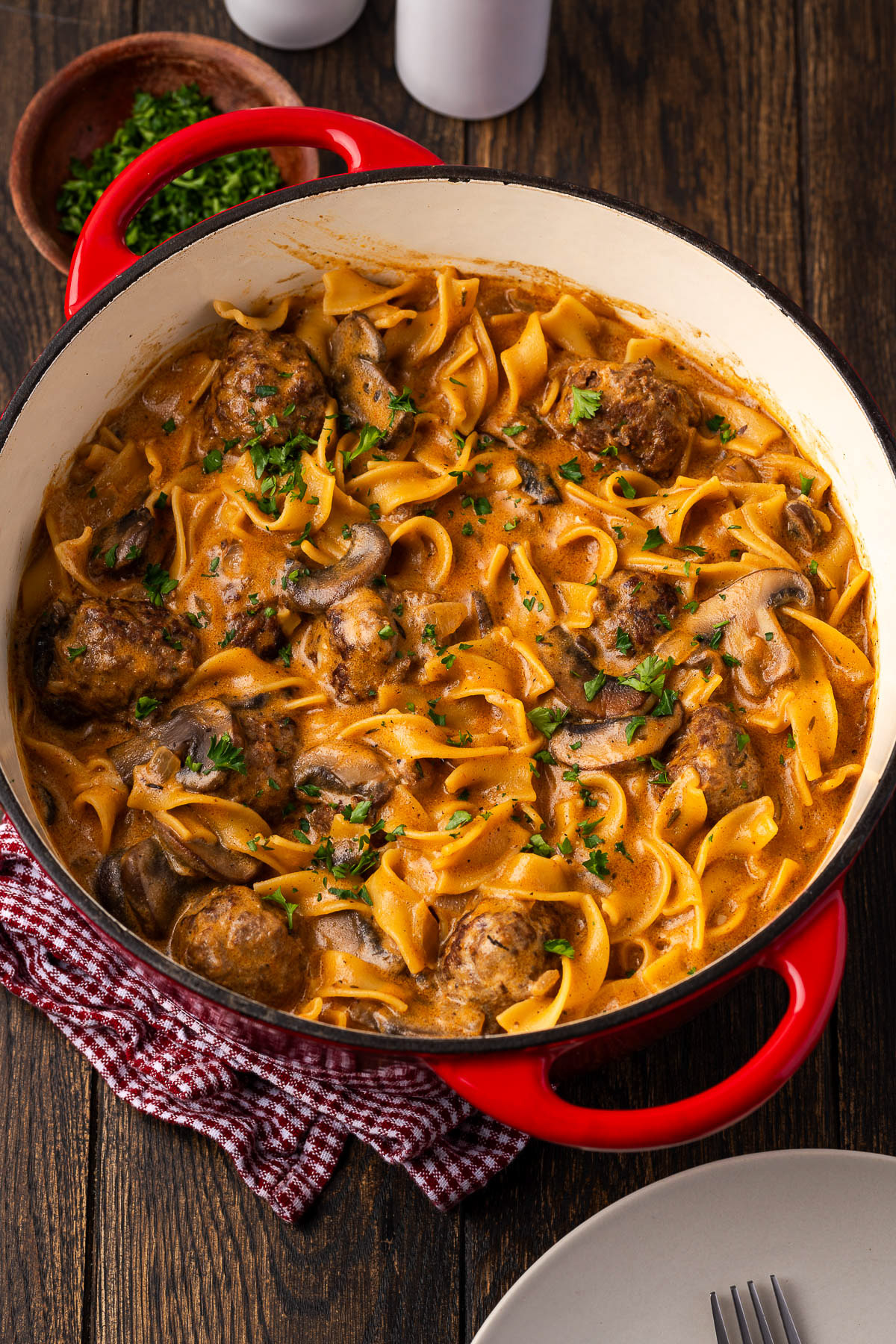 Meatball stroganoff with egg noodles and mushrooms in a red cast iron Dutch oven, plate and parsley beside the pot