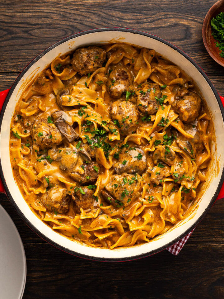 Overhead view of meatball stroganoff in a cast iron Dutch oven with plates, forks and fresh parsley