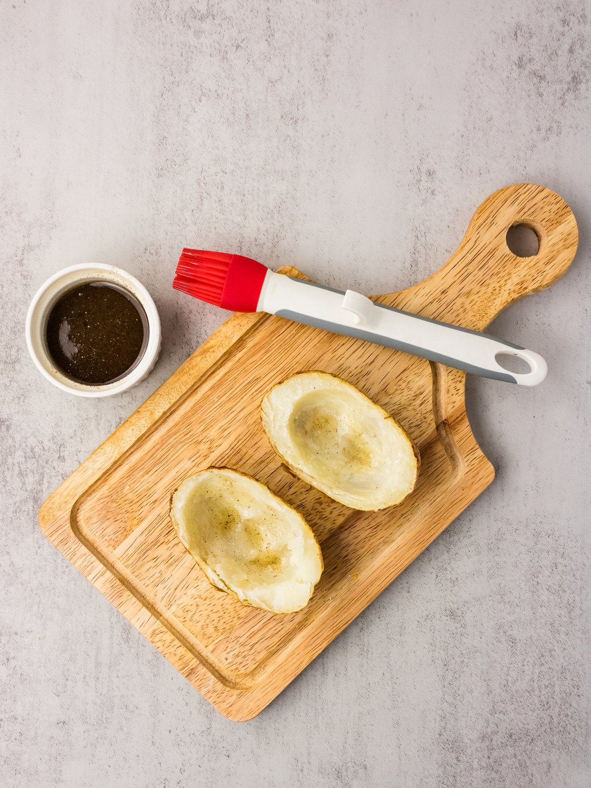 Potato skin shells on a cutting board next to a bowl of bacon grease and a silicone brush