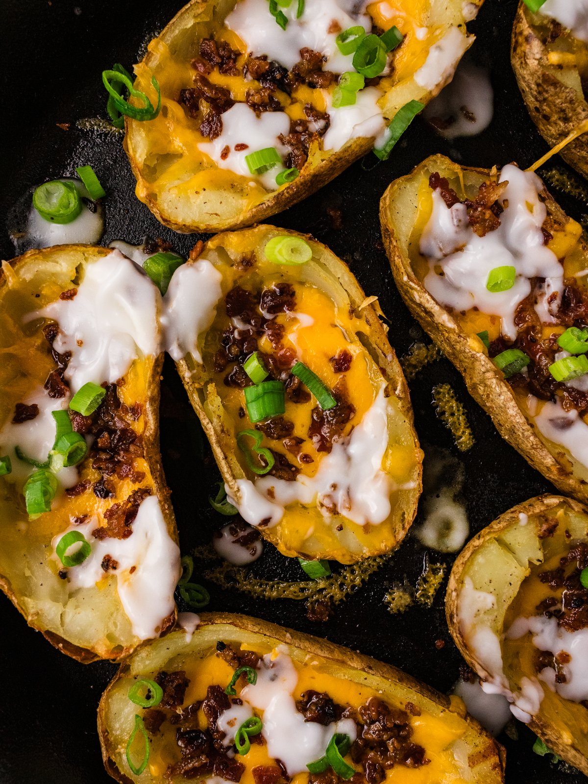 Close up of finished potato skins with melted cheddar, bacon, sour cream and green onions in a cast iron skillet